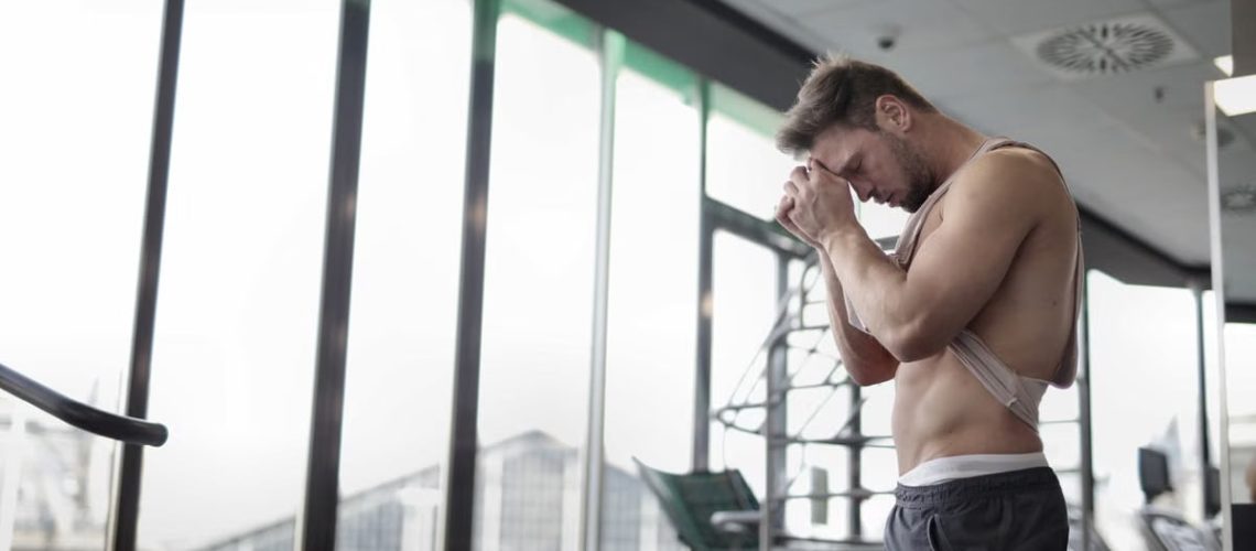 Side view of sweaty muscular male athlete in casual sportswear standing with hands on forehead and resting after intense workout in light fitness center